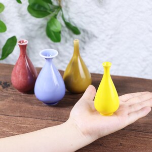 Puede incluir: Una mano sostiene un pequeño jarrón de cerámica amarillo. Hay otros tres jarrones en el fondo, en rojo, azul y dorado. Los jarrones tienen forma de lágrima con cuello estrecho y borde acampanado. Decoración.