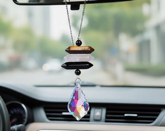 May include: A car charm with a layered design. It features a clear, iridescent crystal pendant at the bottom, with a stack of brown, white, and black crystal-like shapes above. The charm hangs from a silver chain, suspended from a car's rearview mirror.