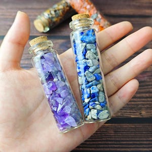 Op de afbeelding: Twee kleine glazen flesjes met kurken stoppen, gevuld met kleurrijke stenen. Eén flesje bevat paarse amethiststenen en de andere blauwe lapis lazuli stenen. De flesjes worden in een hand gehouden.