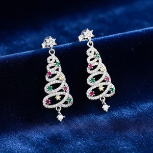 Könnte beinhalten: Ein Paar silberfarbene Weihnachtsbaum-Ohrringe. Jeder Ohrring hat ein Spiraldesign mit klaren und farbigen Edelsteinen, darunter Rot, Grün und Gelb. Die Ohrringe haben einen Steckerverschluss und einen kleinen, hängenden Edelstein am unteren Ende.
