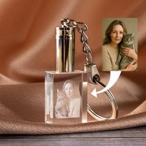 May include: A clear crystal keychain with a silver metal top and chain. The crystal cube features a 3D image of a woman holding a cat. The background is a soft, draped fabric.