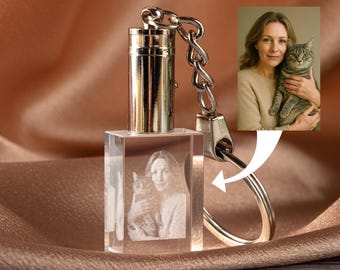 May include: A clear crystal keychain with a silver metal top and chain. The crystal cube features a 3D image of a woman holding a cat. The background is a soft, draped fabric.