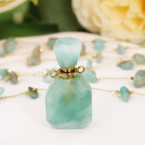 Könnte beinhalten: Eine facettierte, helltürkisfarbene Parfümflasche mit passendem Verschluss. Die Flasche hat einen goldfarbenen Metallkragen. Eine Halskette mit türkisfarbenen Steinen und goldener Kette befindet sich im Hintergrund. Die Flasche steht auf einer weißen Oberfläche.