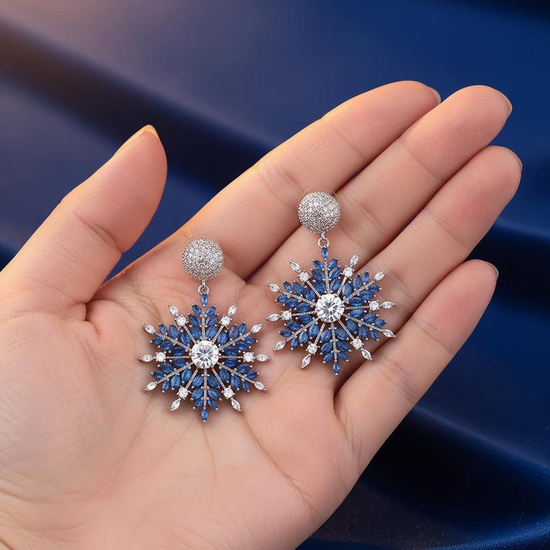 May include: A pair of snowflake-shaped earrings. Each earring features a central clear stone surrounded by blue and white stones, with a small, round, silver-colored stud at the top. The earrings are held in a hand against a dark blue background.