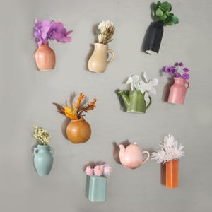 May include: A collection of ten small wall-mounted ceramic vases in various shapes and colors, including peach, cream, black, green, pink, light blue, orange, and sage, each holding a different arrangement of dried or artificial flowers.