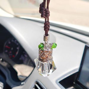 May include: A car air freshener with a brown cord and a decorative design. The freshener has a clear, faceted bottom, a silver filigree midsection, and two green glass beads. The car interior is visible in the background.