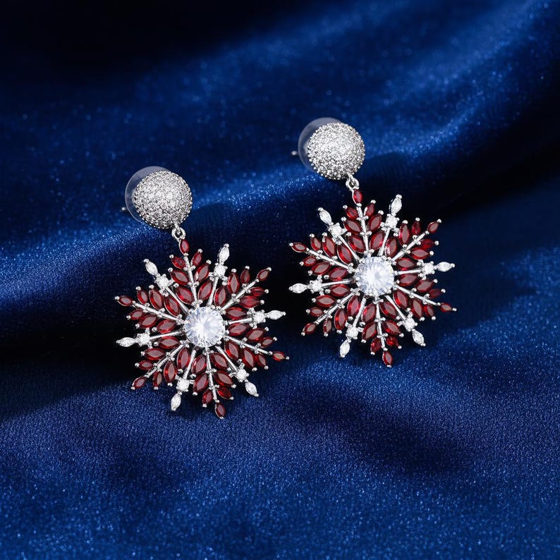May include: A pair of snowflake-shaped earrings. Each earring features a central white gemstone surrounded by red, oval-shaped stones and smaller white accents. The earrings have a silver-colored post and a matching, round, silver-colored top.