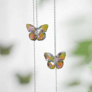 May include: Two iridescent, faceted butterfly-shaped ornaments suspended from silver-toned ball chains. Each butterfly features a central cluster of small, clear crystals and displays shifting hues of yellow, orange, and green across its surface.