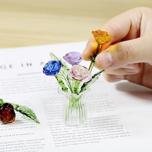 May include: A miniature glass bouquet with blue, pink, purple, and orange roses in a small clear vase. The delicate glass petals and green stems create a charming, handcrafted floral display. The bouquet is a decorative item.