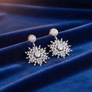 May include: A pair of silver-toned snowflake earrings. Each earring features a round, crystal-studded stud, from which a detailed snowflake design hangs. The snowflake design has a central clear crystal and is surrounded by smaller, leaf-like crystals. The earrings are displayed on a blue velvet surface.