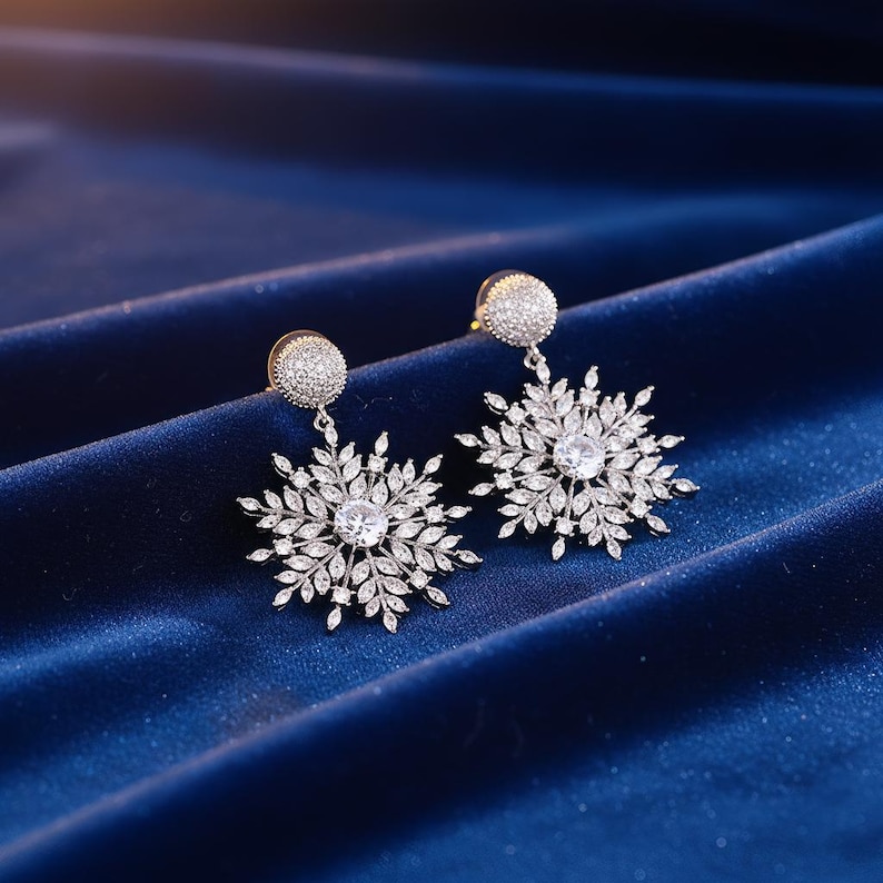 May include: A pair of silver-toned snowflake earrings. Each earring features a round, crystal-studded stud, from which a detailed snowflake design hangs. The snowflake design has a central clear crystal and is surrounded by smaller, leaf-like crystals. The earrings are displayed on a blue velvet surface.