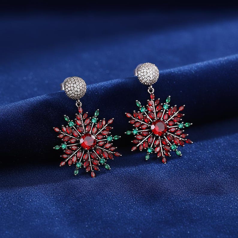May include: A pair of snowflake-shaped earrings with a central red gemstone surrounded by red and green stones. The earrings have a silver-colored post with a small, round, textured design. The background is a dark blue fabric.