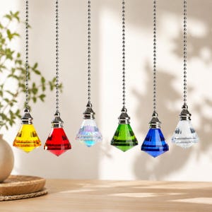 May include: Six faceted crystal ceiling fan pulls hanging from silver-toned metal ball chains. The pendants are shaped like diamonds and come in yellow, red, iridescent, green, blue, and clear glass, each topped with a small silver metal cap.