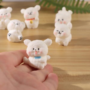 May include: A collection of small white bear figurines with rounded ears, pink cheeks, and black facial features. One figurine is held in the foreground, featuring a light blue collar, while others in the background display variations like a red collar.