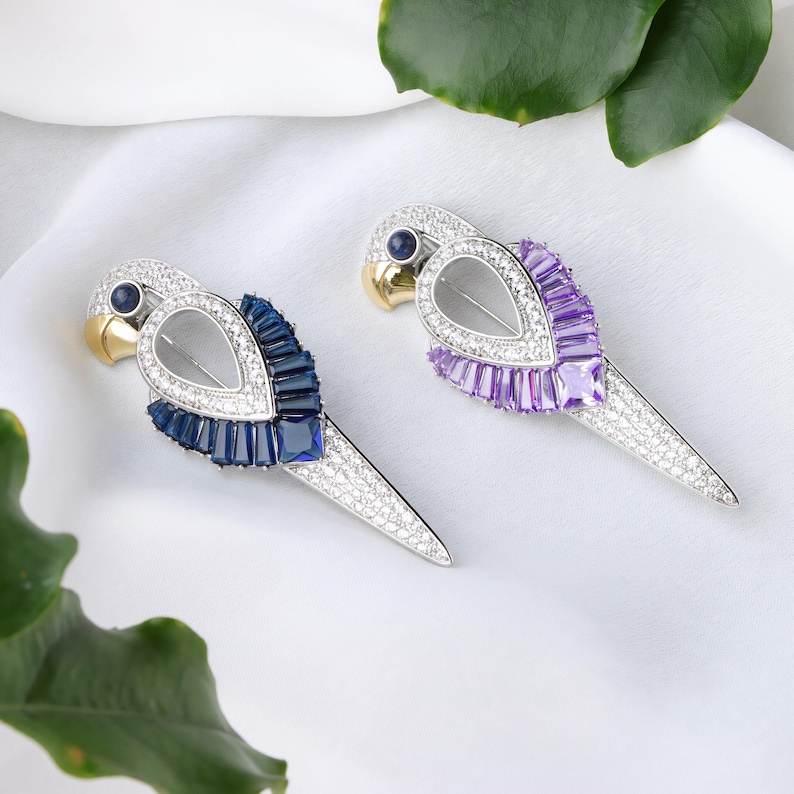 Puede incluir: Dos broches con forma de loro. Un broche presenta piedras preciosas azules y transparentes, con un pico dorado y un ojo de piedra preciosa azul. El otro broche tiene piedras preciosas moradas y transparentes, un pico dorado y un ojo de piedra preciosa azul.