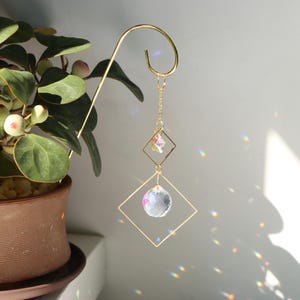 Puede incluir: Adorno colgante de metal dorado con una bola de cristal transparente y una estrella de cristal más pequeña. El adorno está suspendido de un gancho curvo, con un marco cuadrado alrededor de cada cristal. Un planta en maceta está en el fondo.