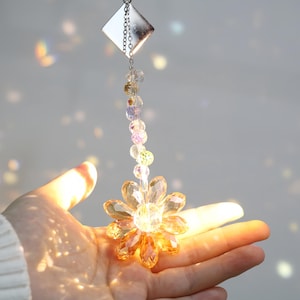 Op de afbeelding: Een decoratieve zonnevanger met een hanger van kristal in de vorm van een madeliefje. De hanger is gemaakt van facetgeslepen, amberkleurig glas en hangt aan een ketting van kleurrijke kralen en een zilverkleurige metalen ketting. De zonnevanger wordt in een hand gehouden.