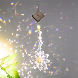 May include: A clear crystal daisy suncatcher with a yellow centre and faceted petals hangs from a chain. A silver diamond-shaped charm above it reads "Happy birthday, Mom!". The suncatcher is adorned with colourful beads.