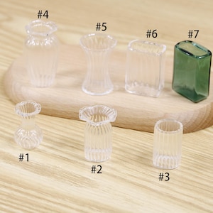 Puede incluir: Una colección de jarrones de vidrio en miniatura de varias formas y tamaños. Los jarrones son transparentes, con diseños acanalados y estriados. Un jarrón tiene un tinte verde. Los jarrones están dispuestos sobre una tabla de cortar de madera.