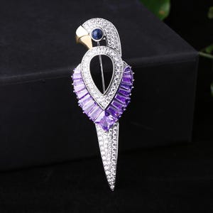 Puede incluir: Broche con forma de loro con cuerpo de metal plateado, adornado con cristales transparentes y morados. El loro tiene un pico dorado y un ojo de gema azul. Accesorio decorativo.
