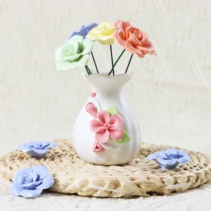 May include: A white ceramic vase with a pink floral design holds a bouquet of colorful artificial roses. The roses are in shades of green, yellow, orange, and blue. Two blue roses are placed on a woven mat.