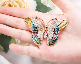 May include: A gold butterfly brooch with vibrant enamel wings and crystal embellishments. The brooch has a central pink gem and small red gems on the antennae. The wings are a mosaic of colours including blue, green, orange, and pink, creating a striking design.