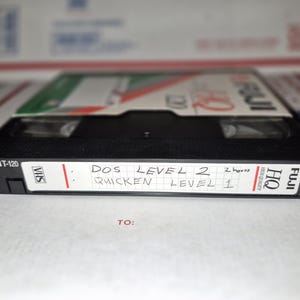 May include: A black VHS tape labeled FUJI-120 with the text "DOS LEVEL 2" and "QUICKEN LEVEL 1" written on the label. The tape is resting on a white surface with a partially visible box in the background. The tape also has the text "HQ HIGH QUALITY" on the label.