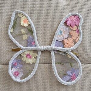 May include: Delicate butterfly wings with a white trim and a gold elastic band. The wings are adorned with embroidered flowers in shades of pink, purple, yellow, and orange, creating a whimsical and enchanting accessory.
