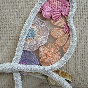 May include: Close-up of a decorative floral applique. The applique features embroidered flowers in shades of pink, peach, and purple, outlined with white trim. A gold ribbon is visible. The design is set against a sheer fabric.