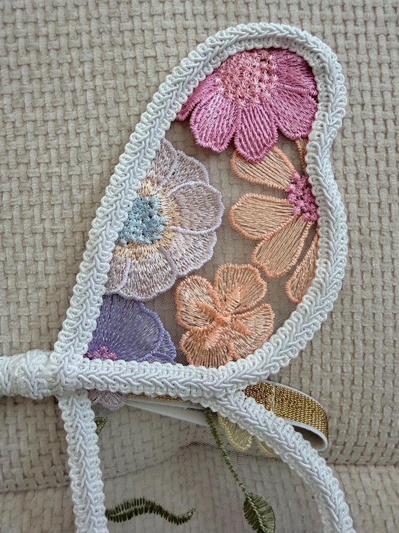 May include: Close-up of a decorative floral applique. The applique features embroidered flowers in shades of pink, peach, and purple, outlined with white trim. A gold ribbon is visible. The design is set against a sheer fabric.