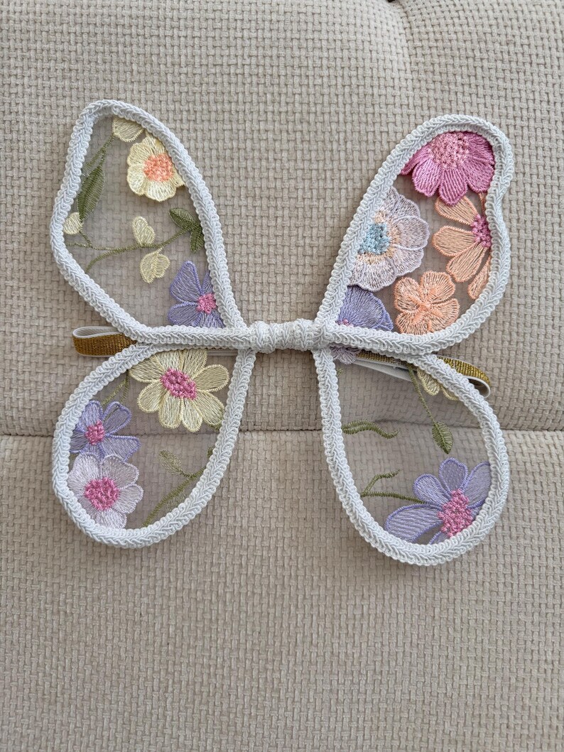 May include: Delicate butterfly wings with a white trim and a gold elastic band. The wings are adorned with embroidered flowers in shades of pink, purple, yellow, and orange, creating a whimsical and enchanting accessory.