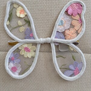May include: Embroidered butterfly wings with a white trim and a gold elastic band. The wings are filled with colorful floral embroidery in shades of pink, yellow, and purple, creating a whimsical accessory.