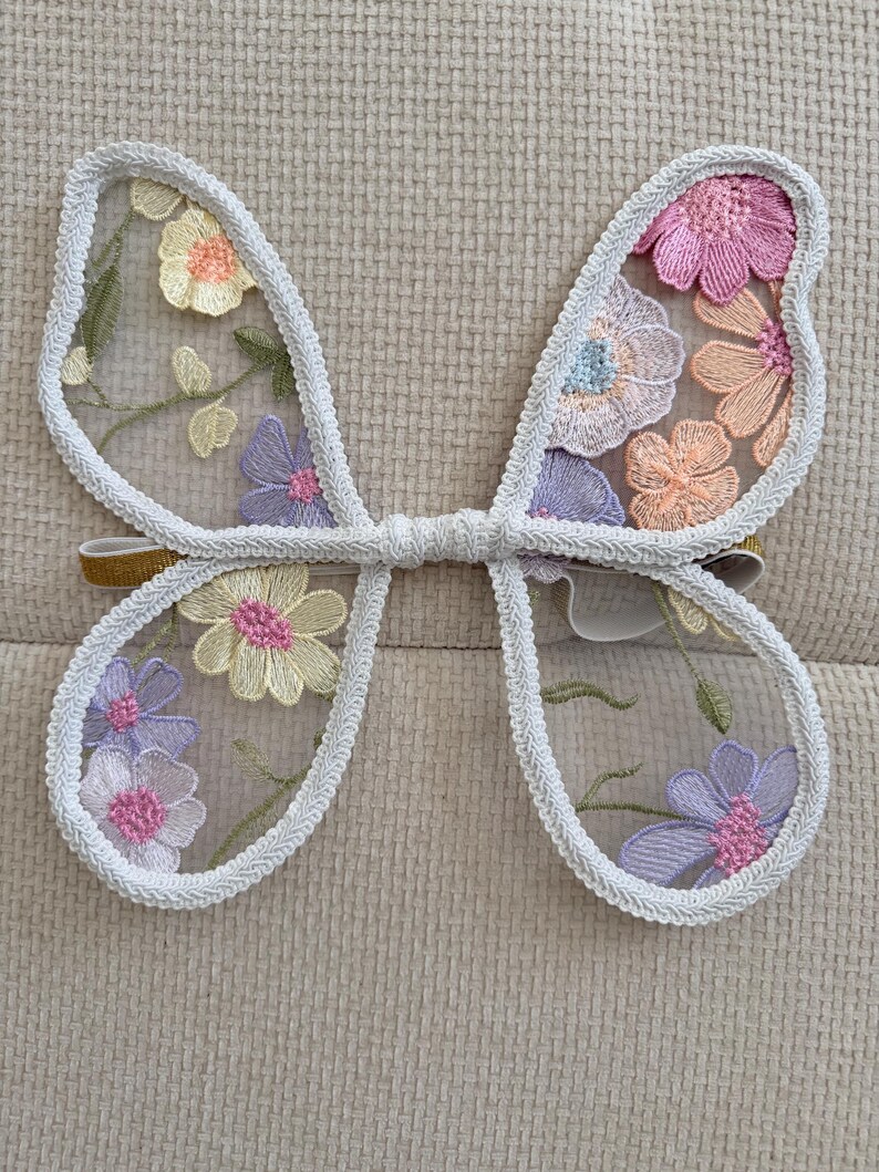 May include: Embroidered butterfly wings with a white trim and a gold elastic band. The wings are filled with colorful floral embroidery in shades of pink, yellow, and purple, creating a whimsical accessory.