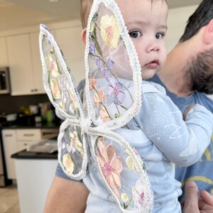 May include: A baby wearing light blue pajamas and a pair of sheer butterfly wings. The wings are white with embroidered flowers in shades of pink, yellow, and purple. The baby is being held by an adult.