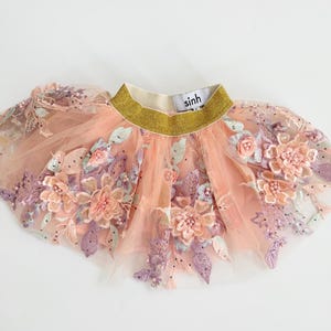 May include: A peach-colored tulle skirt with intricate floral embroidery in shades of pink, lavender, and green. The waistband is gold with the brand name "sinh" in white. The skirt is designed for children.