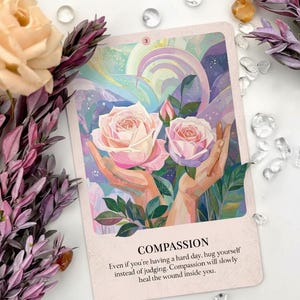 May include: An oracle card with the word "COMPASSION" and the number "3". The card features a painting of two pink roses held by hands, with a soft, pastel background. Text on the card encourages self-compassion. Decorative flowers and crystals surround the card.