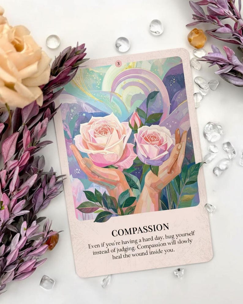 May include: An oracle card with the word "COMPASSION" and the number "3". The card features a painting of two pink roses held by hands, with a soft, pastel background. Text on the card encourages self-compassion. Decorative flowers and crystals surround the card.