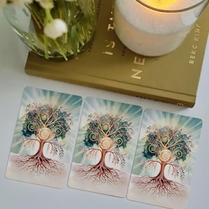 May include: Three tarot cards featuring a tree of life design with intricate branches and roots. The cards are on a white surface, next to a lit candle in a glass jar and a book. The tree has a spiral design in the center.