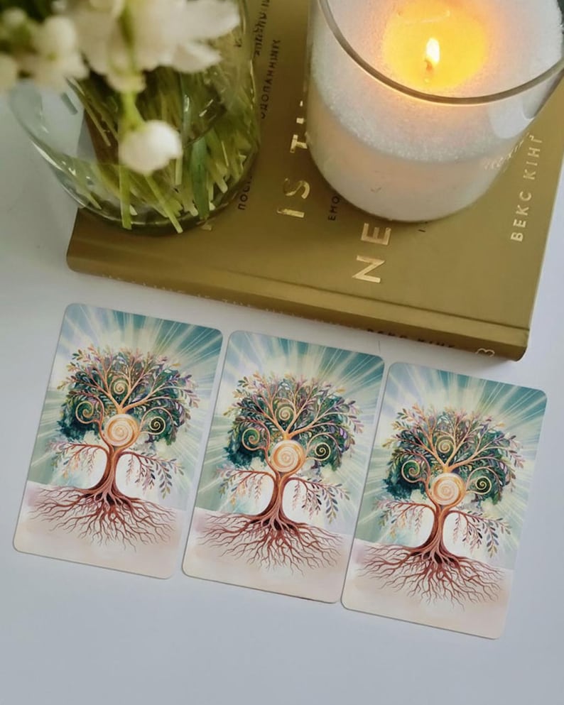 May include: Three tarot cards featuring a tree of life design with intricate branches and roots. The cards are on a white surface, next to a lit candle in a glass jar and a book. The tree has a spiral design in the center.