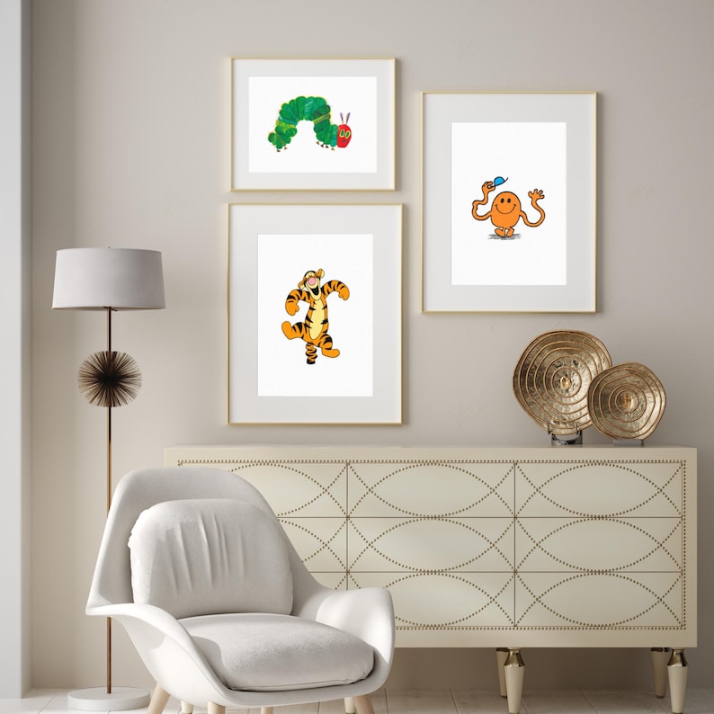 May include: Three framed prints on a white wall above a white dresser with a gold metal decorative stand. The prints feature a green caterpillar, a cartoon tiger, and a yellow smiley face with arms and legs.