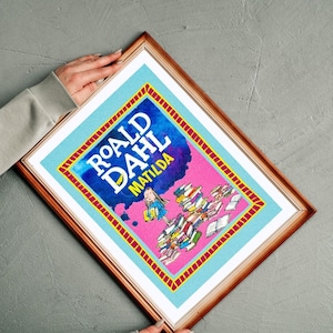 May include: Framed print of the book cover for Roald Dahl's "Matilda." The design features a pink backdrop with the title in blue and an illustration of a girl with books. The frame is a warm brown colour.