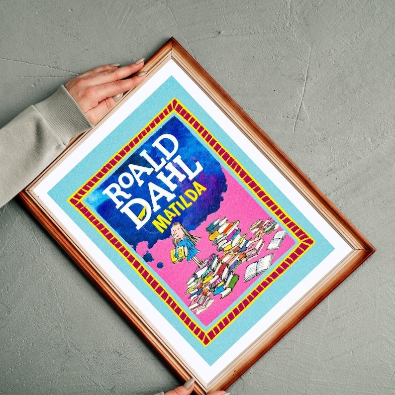 May include: Framed print of the book cover for Roald Dahl's "Matilda." The design features a pink backdrop with the title in blue and an illustration of a girl with books. The frame is a warm brown colour.