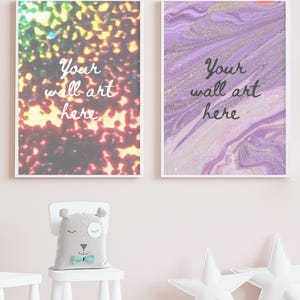 May include: Two framed wall art prints with the text "Your wall art here" on a pink wall. The prints are in a modern style with abstract designs. One print features a purple and gold marble design, and the other features a colourful abstract design. The prints are hanging above a white chair with a grey stuffed animal sitting on it.