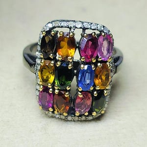 May include: A silver ring with a rectangular cluster of colorful gemstones. The gemstones are in shades of yellow, orange, pink, blue, and green. The ring is set with small, clear stones around the cluster.