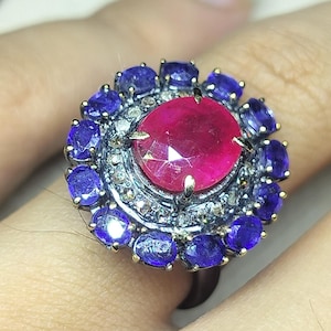 Puede incluir: Un anillo de plata con una gran gema central de color rubí, rodeada de zafiros azules oscuros más pequeños. El anillo tiene un diseño floral y está sobre un fondo claro.