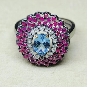 May include: A silver ring with a floral design. The ring features a central oval-shaped light blue gemstone, surrounded by a halo of small white and red gemstones. The ring is set against a neutral background.