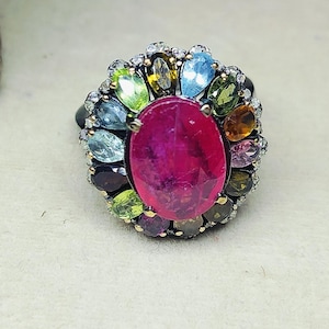 Può includere: Un anello d'argento con una grande gemma magenta ovale al centro, circondata da una serie di gemme più piccole e colorate. Il design dell'anello comprende varie tonalità, come verde, blu e giallo, creando un gioiello vibrante.