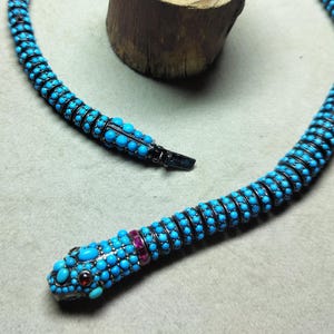 May include: A turquoise beaded snake necklace with a black clasp. The snake's head features red and turquoise accents. The necklace is coiled near a small, round wooden log.