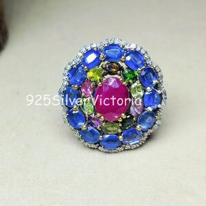 Op de afbeelding: Een ring met een grote ovale roze middensteen omringd door een cluster van kleinere ovale blauwe stenen, afgewisseld met kleine groene en roze edelstenen, gezet in een zilveren frame met de tekst 925SilverVictoria zichtbaar op het oppervlak.