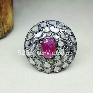 May include: A circular ring featuring a central oval-shaped pinkish-red gemstone surrounded by numerous small, irregular, translucent white stones set in a dark, oxidised metal frame with the text 925SilverVictoria visible on the surface.